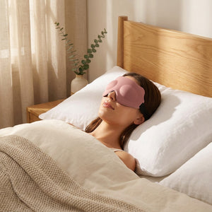 Pink 3D memory foam sleep mask resting on white linen pillow in a serene golden-hour bedroom