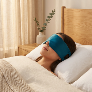 Peacock blue silk sleep mask resting on white linen pillow in a serene golden-hour bedroom